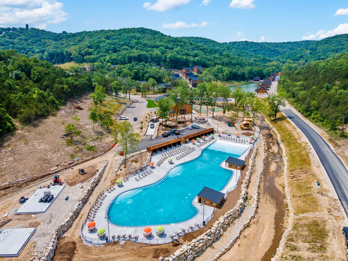 A spacious outdoor swimming pool surrounded by lounge chairs and wooden structures, set against a backdrop of lush green hills. The setting is lively yet tranquil, ideal for sunbathing, swimming, and social summer gatherings.