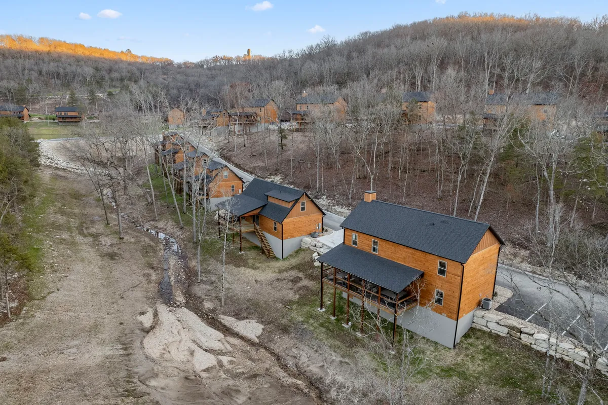 A wide view of several wooden cabins nestled among trees, with a winding road and a tranquil stream running alongside. The natural surroundings are calm and inviting, perfect for family gatherings and nature exploration.