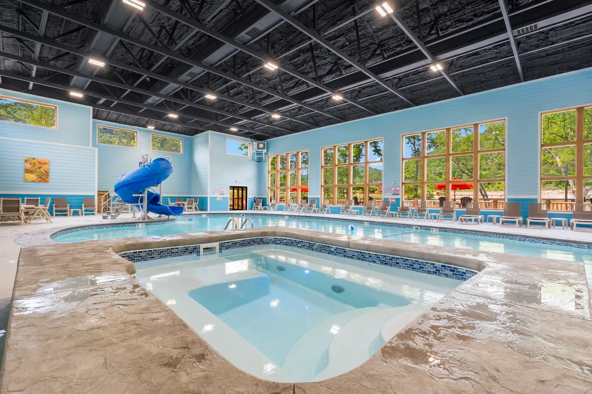 A vibrant indoor pool area featuring a water slide, jacuzzi, and expansive windows that let in plenty of natural light. Families and children are seen enjoying the pool and slide, creating an atmosphere of fun and relaxation.