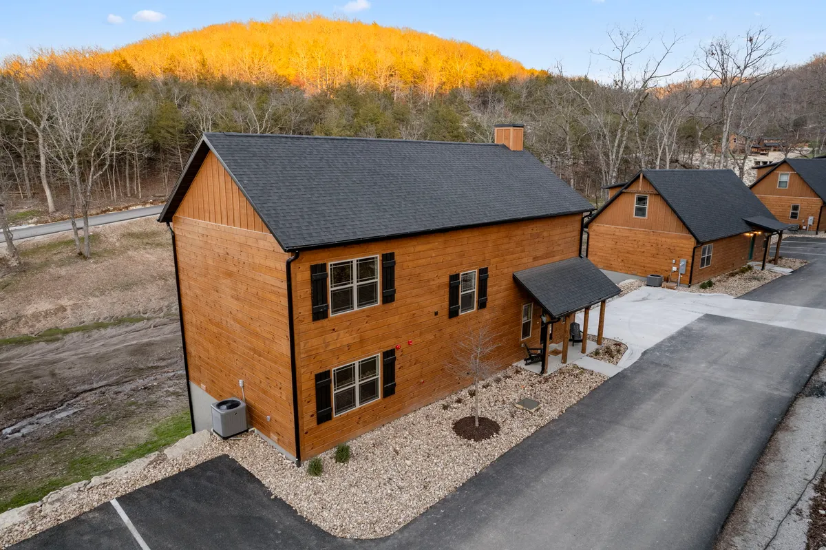 A welcoming view of the cabin's wooden exterior, set against a landscaped yard with a backdrop of mountain views. The scene features a porch, driveway, and hints at the inviting natural surroundings.