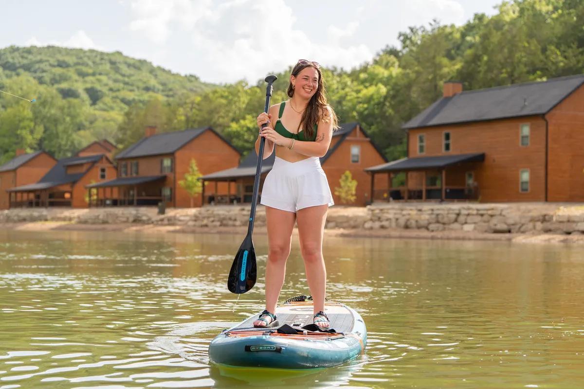 A woman glides across calm water on a paddleboard, with cozy cabins and green hills visible in the background. The tranquil lake setting beckons outdoor recreation and family fun.