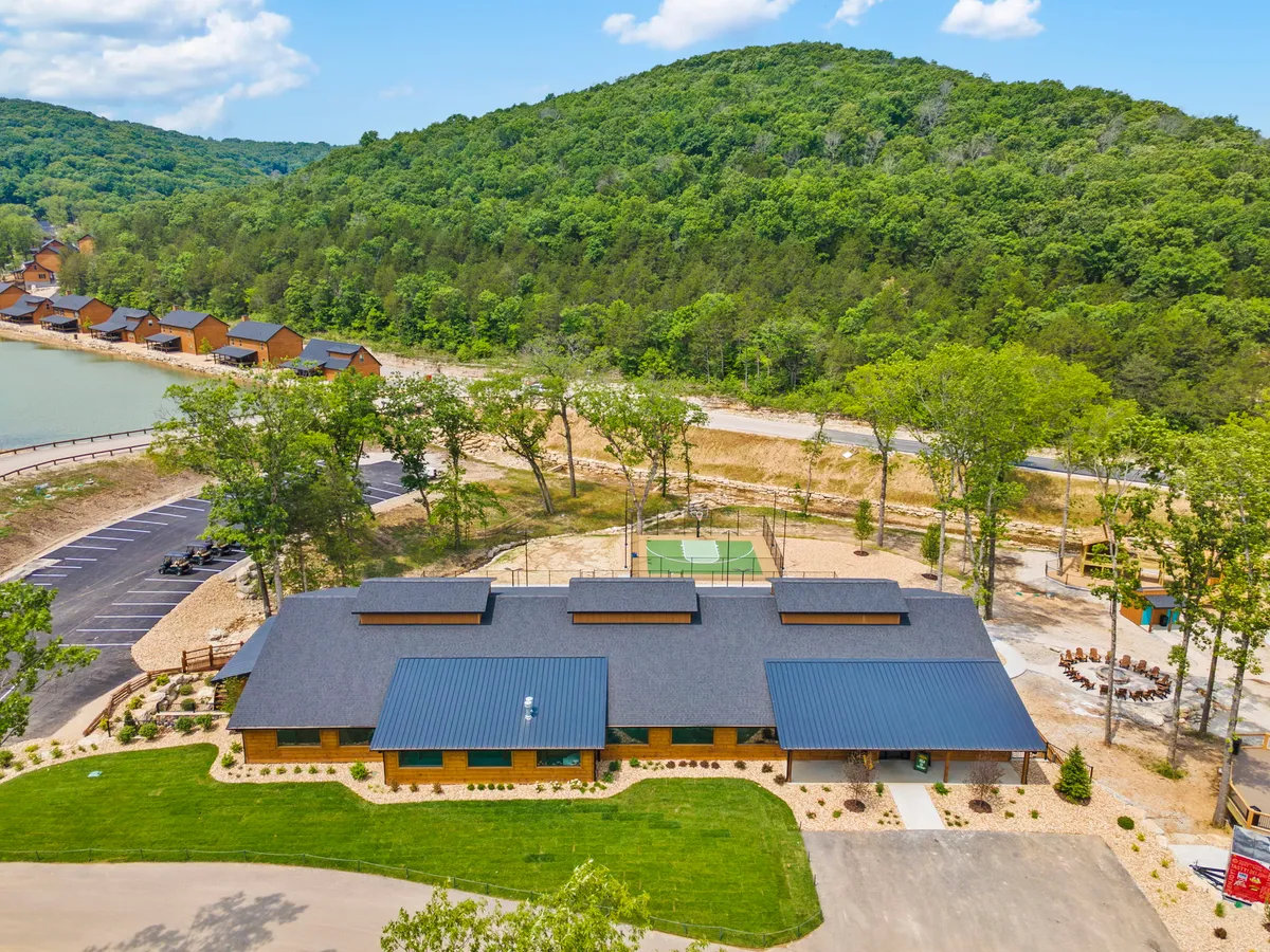 A panoramic exterior shot of the main building with surrounding landscape, recreational facilities, and a visible parking area. The setting suggests easy access to outdoor sports, hiking, boating, and family gatherings amid scenic views.