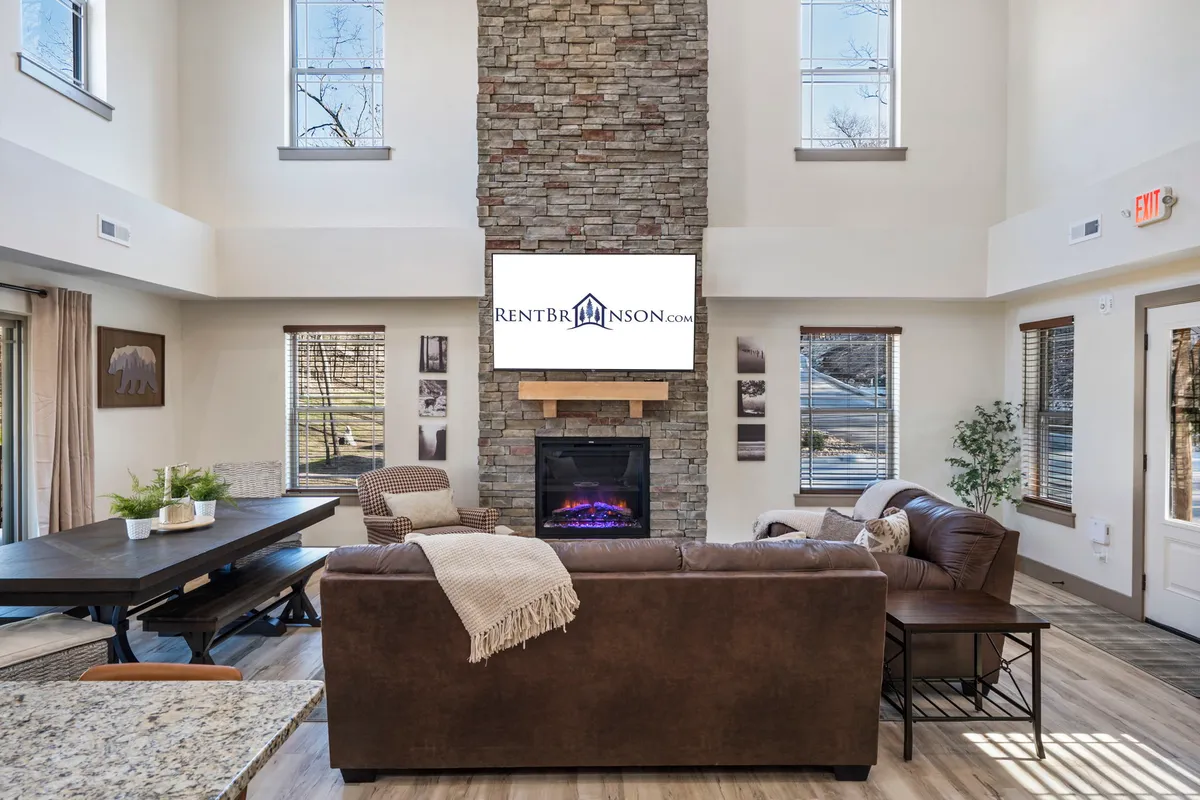 The image shows a warm, inviting interior featuring a rustic stone fireplace as the central focal point, a large TV above the mantel, a spacious dining table ideal for gatherings, and a plush sofa designed for comfort. The ambiance suggests a perfect setting for relaxing evenings, family movie nights, and memorable celebrations.