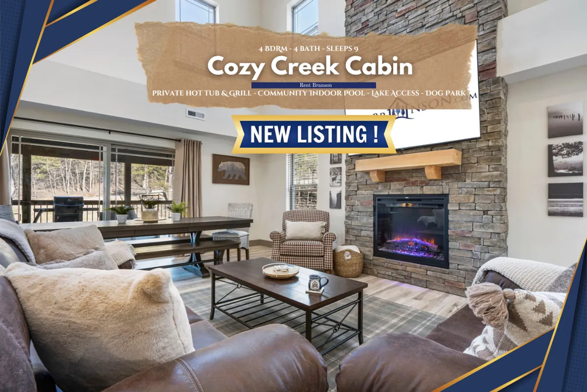 A warm and inviting living/dining area featuring a stone fireplace, comfortable seating, a large wooden dining table, and expansive windows that fill the room with natural light. The décor blends rustic charm with modern touches, and the view through the windows showcases surrounding woodlands.