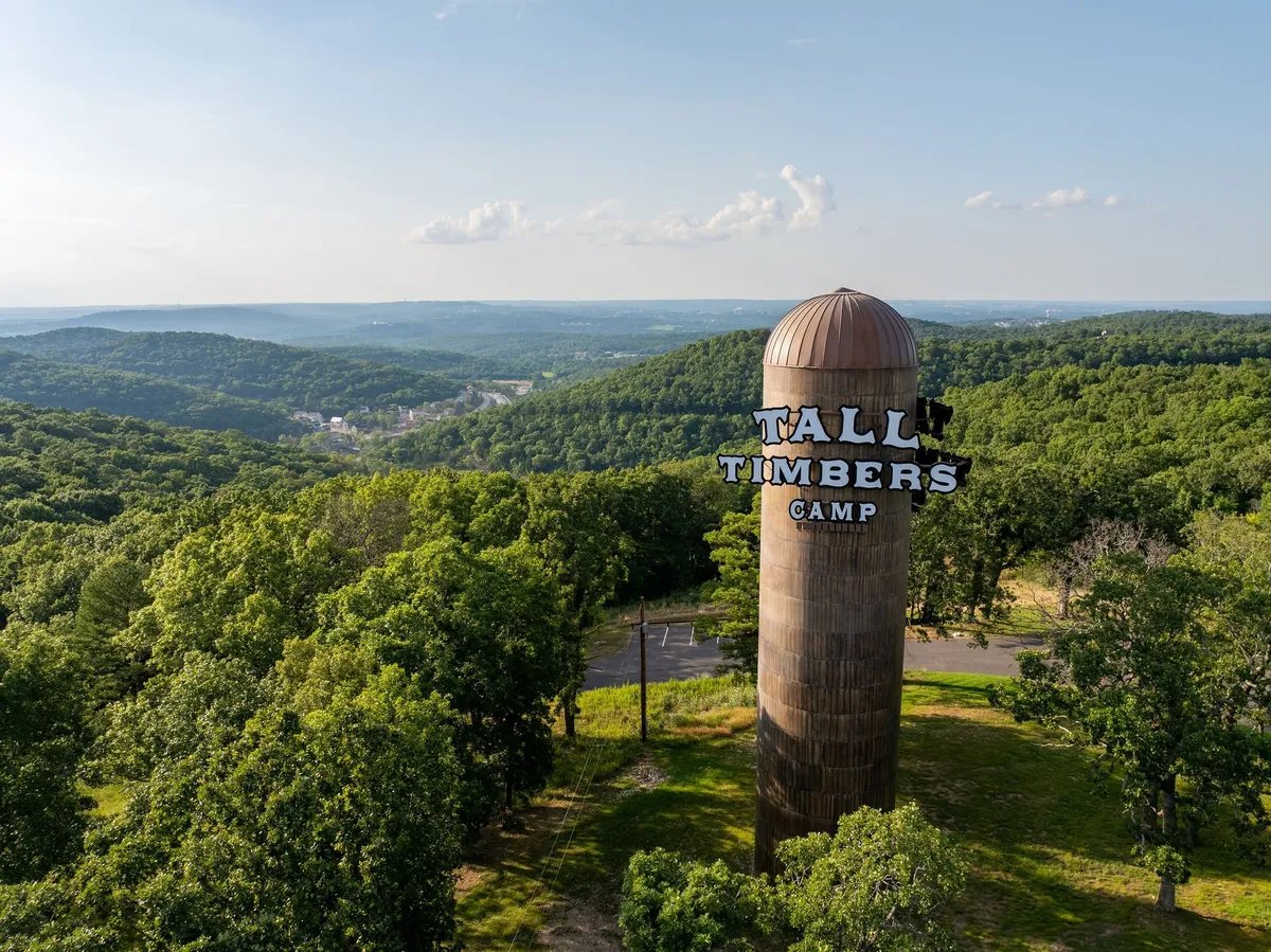 A tall water tower emblazoned with 'Tall Timbers Camp' rises above lush green trees, framed by distant hills. The scene evokes a sense of arrival and adventure, with the camp's natural setting on full display.