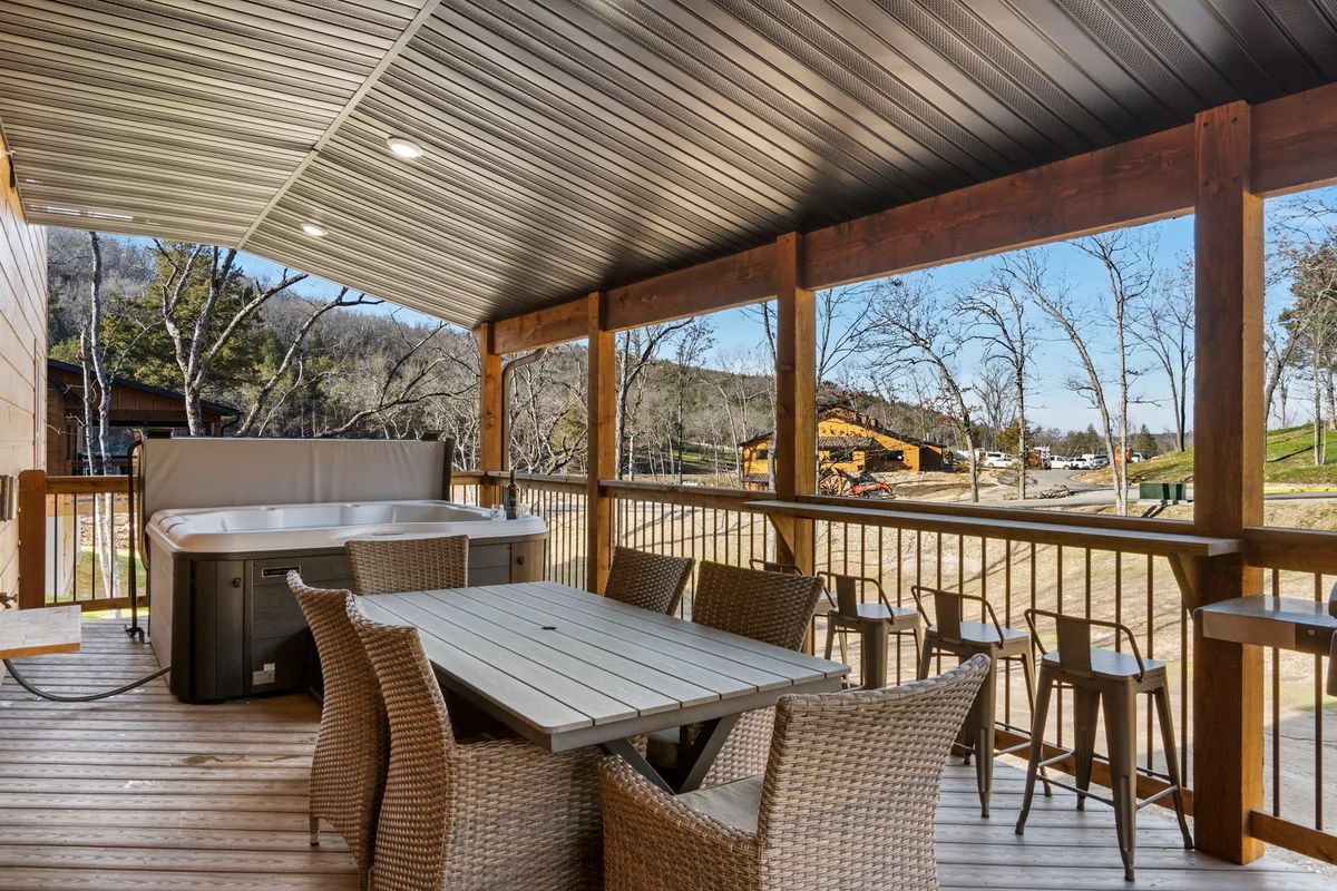 An inviting outdoor deck featuring a dining table and a hot tub with expansive views of rolling hills and treetops. The space is set for socializing, relaxing in the hot tub, and enjoying meals with scenic vistas, perfect for gatherings of all ages.